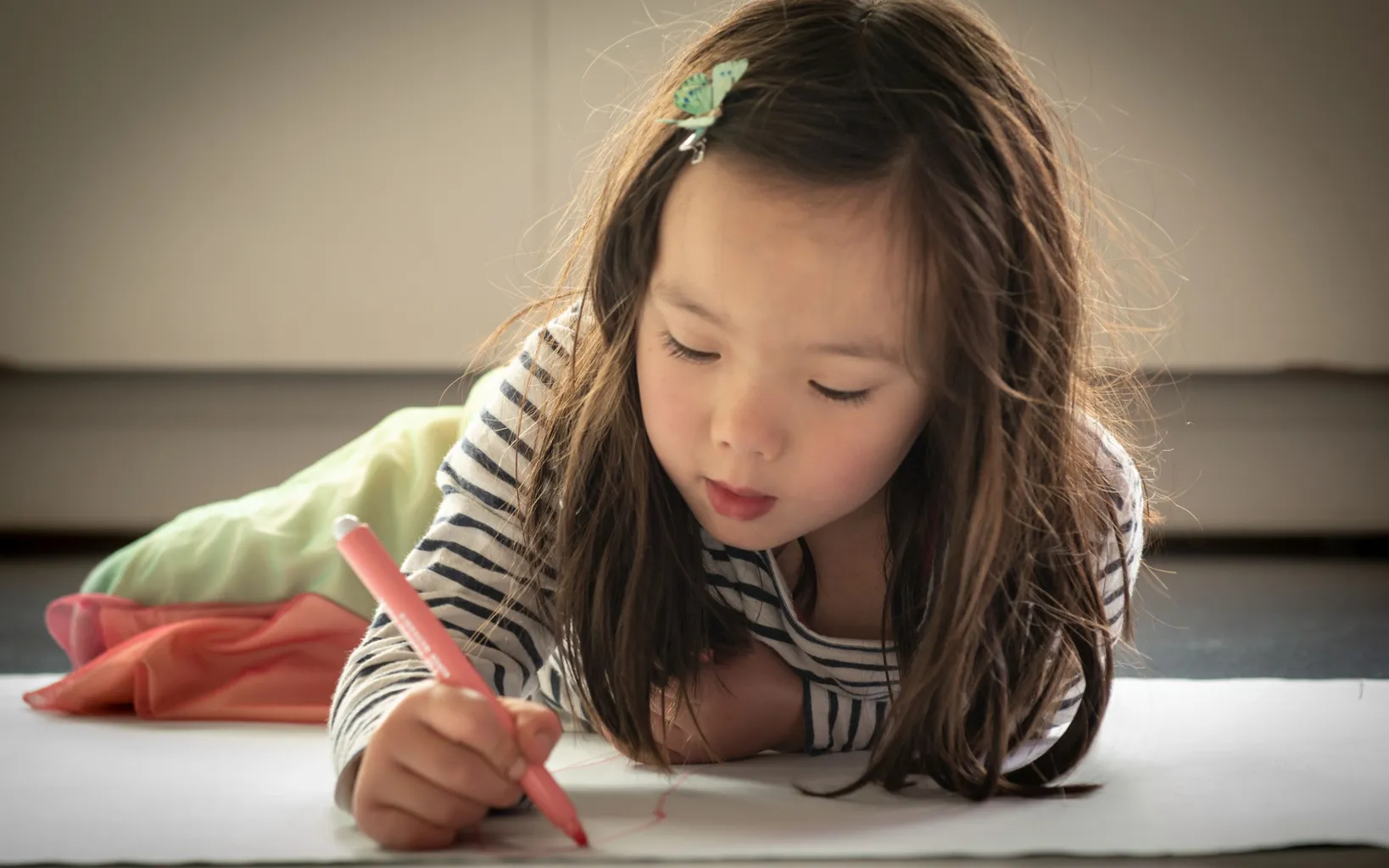 Young girl lying on her stomach and drawing carefully on a large sheet of paper with a pink crayon.