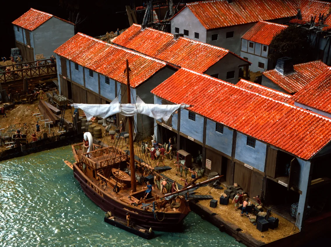 Detailed model of a dockside scene with a ship moored beside several buildings with red-tiled roofs. Numerous small figures populate the area, engaging in various activities.