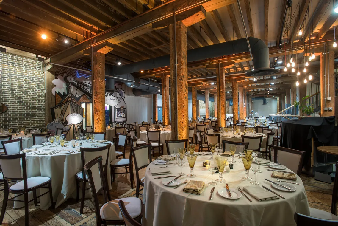 Spacious restaurant with wooden pillars and beams, decorated with round tables set for dining. Dim lighting creates an inviting atmosphere.