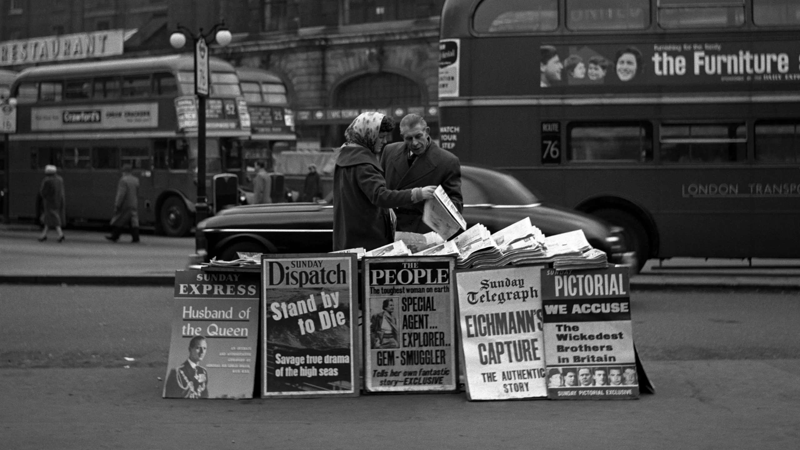 photograph, pigment print. A woman newspaper vendor wearing a headscarf passes a newspaper to a male customer.