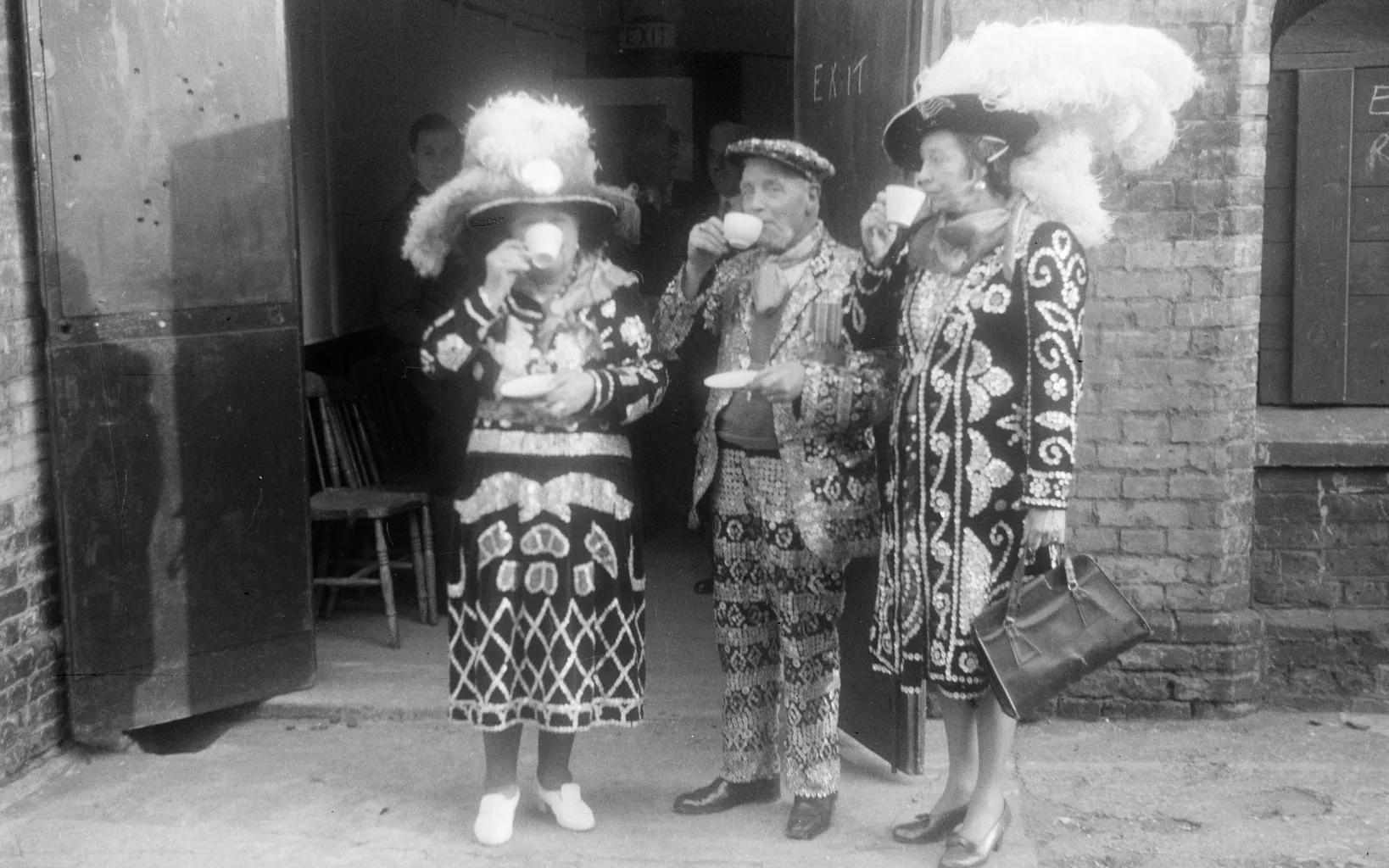 Three women in elaborate, traditional costumes and feathered hats standing outside a building, one eating and another drinking from a cup.