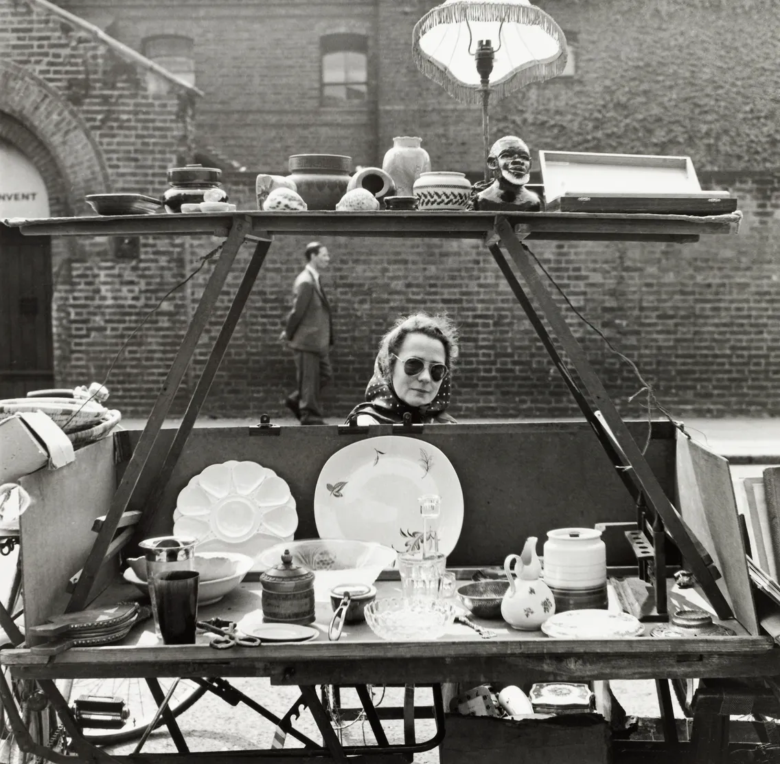 A woman sits behind a makeshift outdoor stall displaying various second-hand goods, including dishes and decorative items, with a brick building in the background.