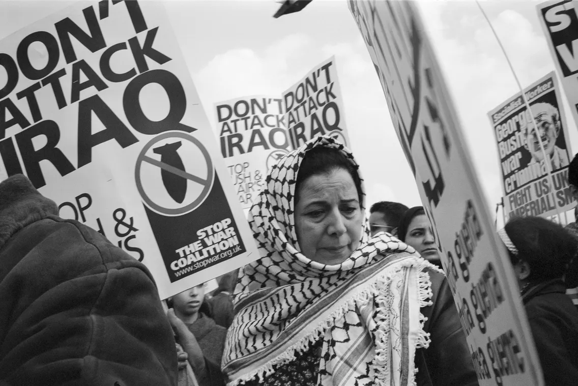 A woman in a headscarf stands solemnly at a protest against military action in iraq, surrounded by placards with peace symbols and anti-war messages.