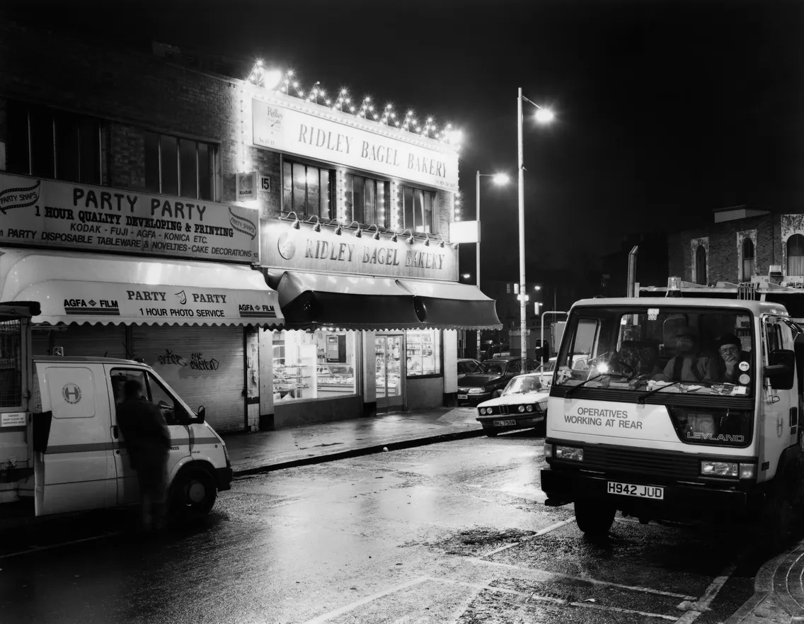 Nighttime street view featuring ridley bagel bakery with illuminated signs, a parked leyland truck, and wet pavement reflecting lights.