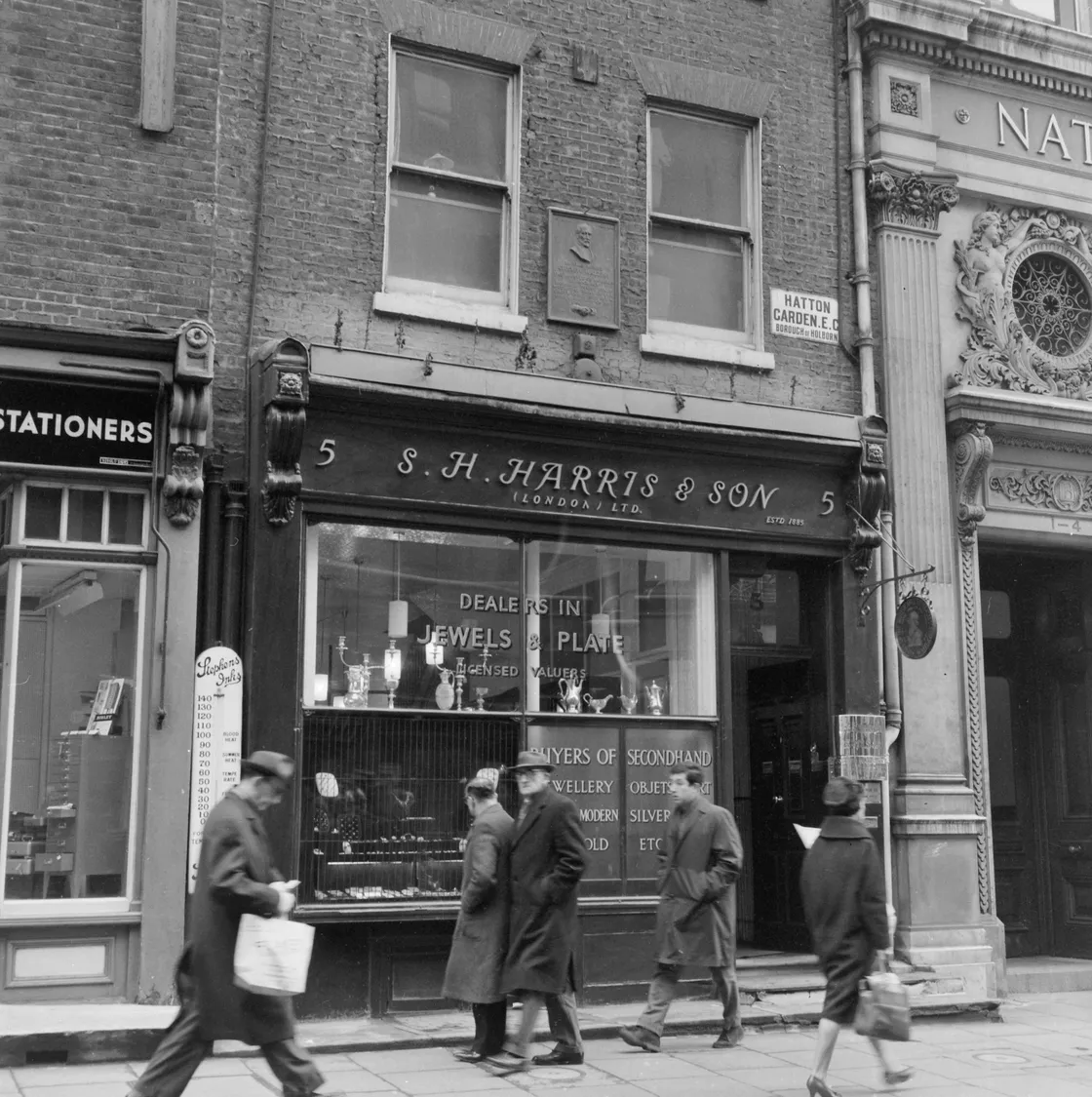 Black and white photo of people walking past vintage shops including a jeweler and stationer on a city street.
