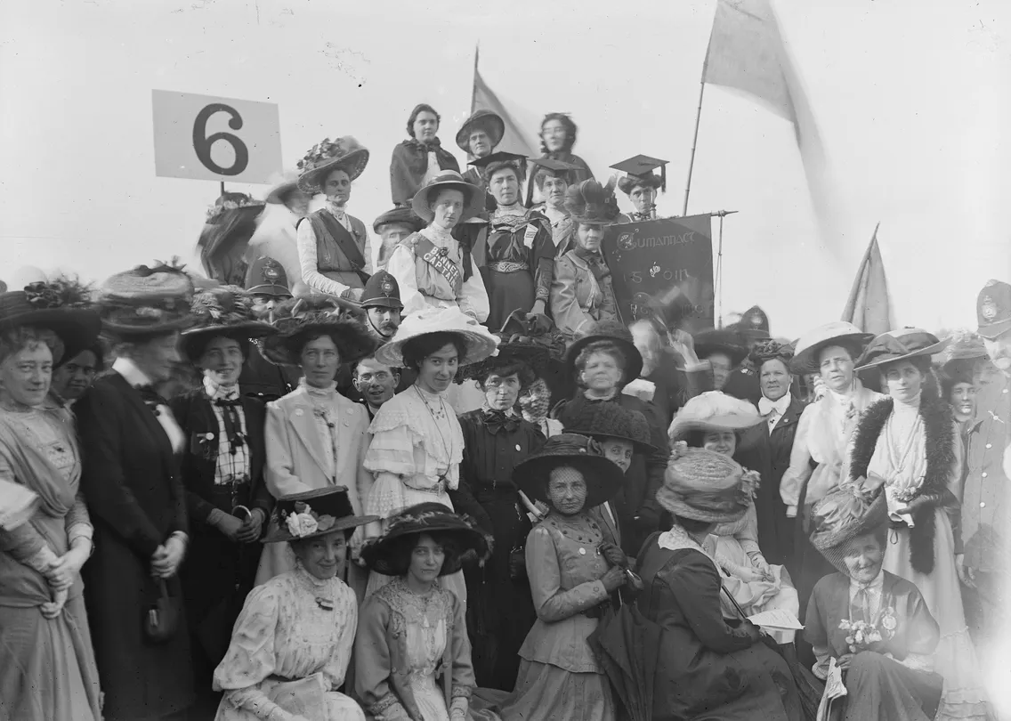 Group of women in early 20th-century attire gathered at an outdoor event, holding banners and flags, with a sign numbered "6" in the background.