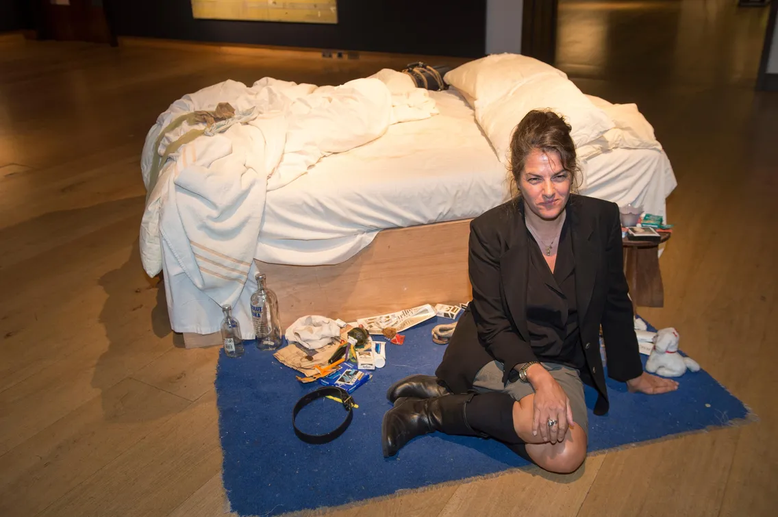 A person sitting on a blue rug next to a cluttered bed in an indoor setting. The bed and surroundings are scattered with various items, including bottles and papers.