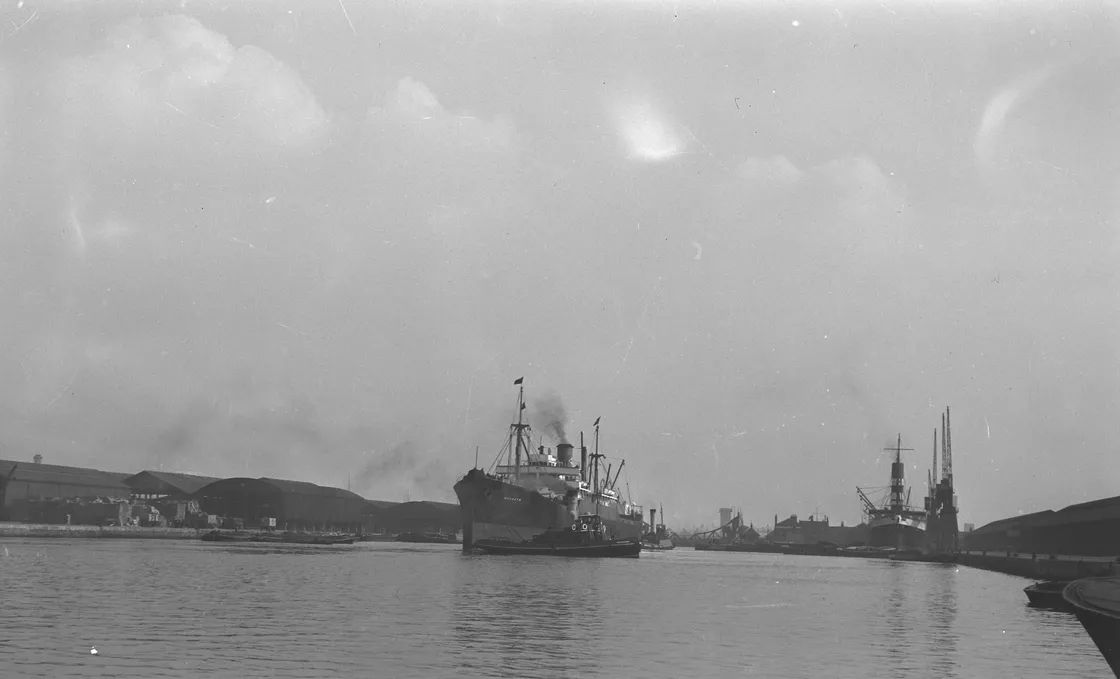 Black and white photo of several large cargo ships docked at an industrial harbour, with warehouses in the background under a cloudy sky.