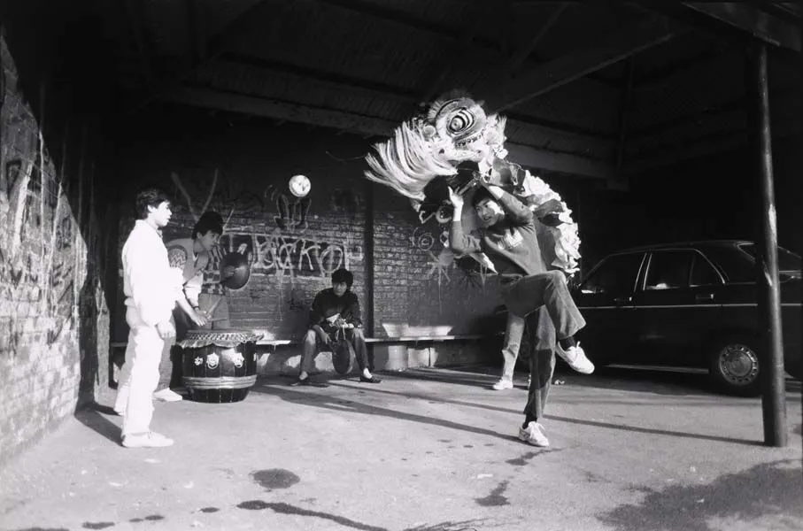 A man performs a dynamic martial arts kick in the air while others watch in an urban, graffiti-covered setting.