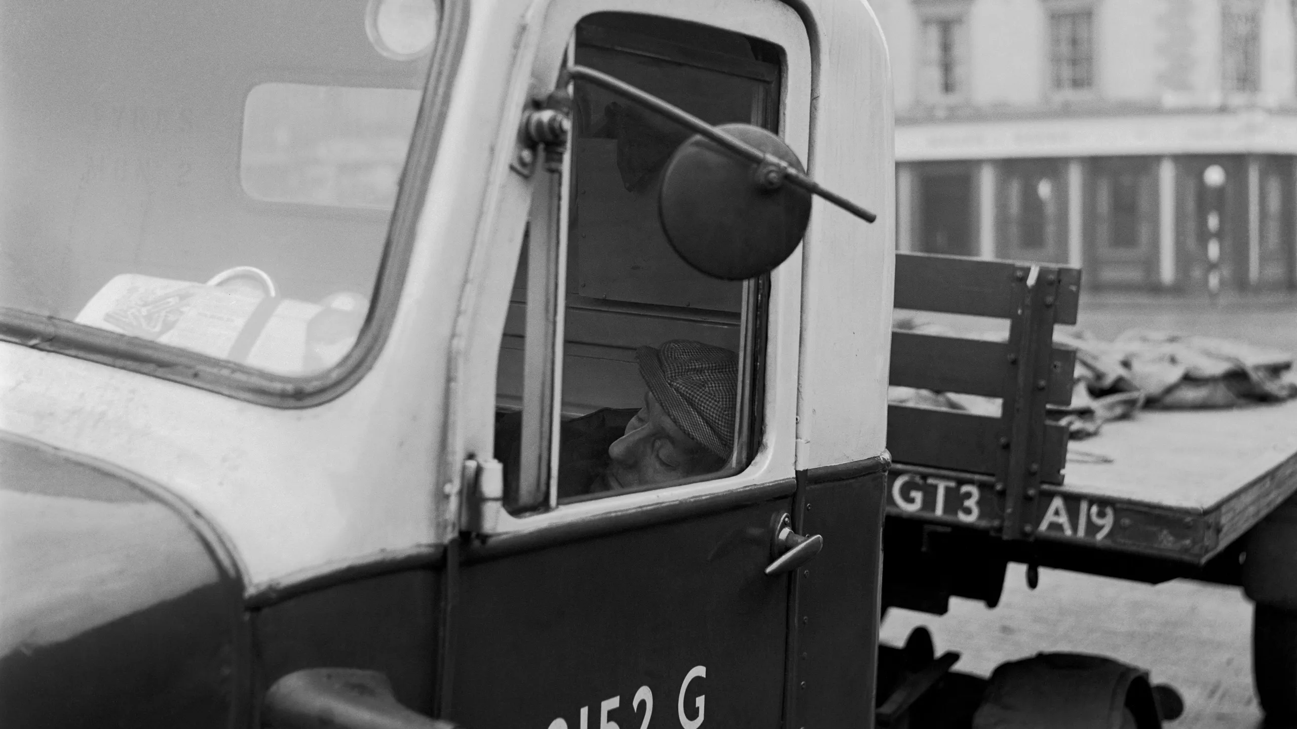 photograph, pigment print. A truck driver, wearing a cap, asleep in the cab of his vehicle. A folded newspaper is visible through the windscreen. On the back of the truck are several empty bags or sacks.