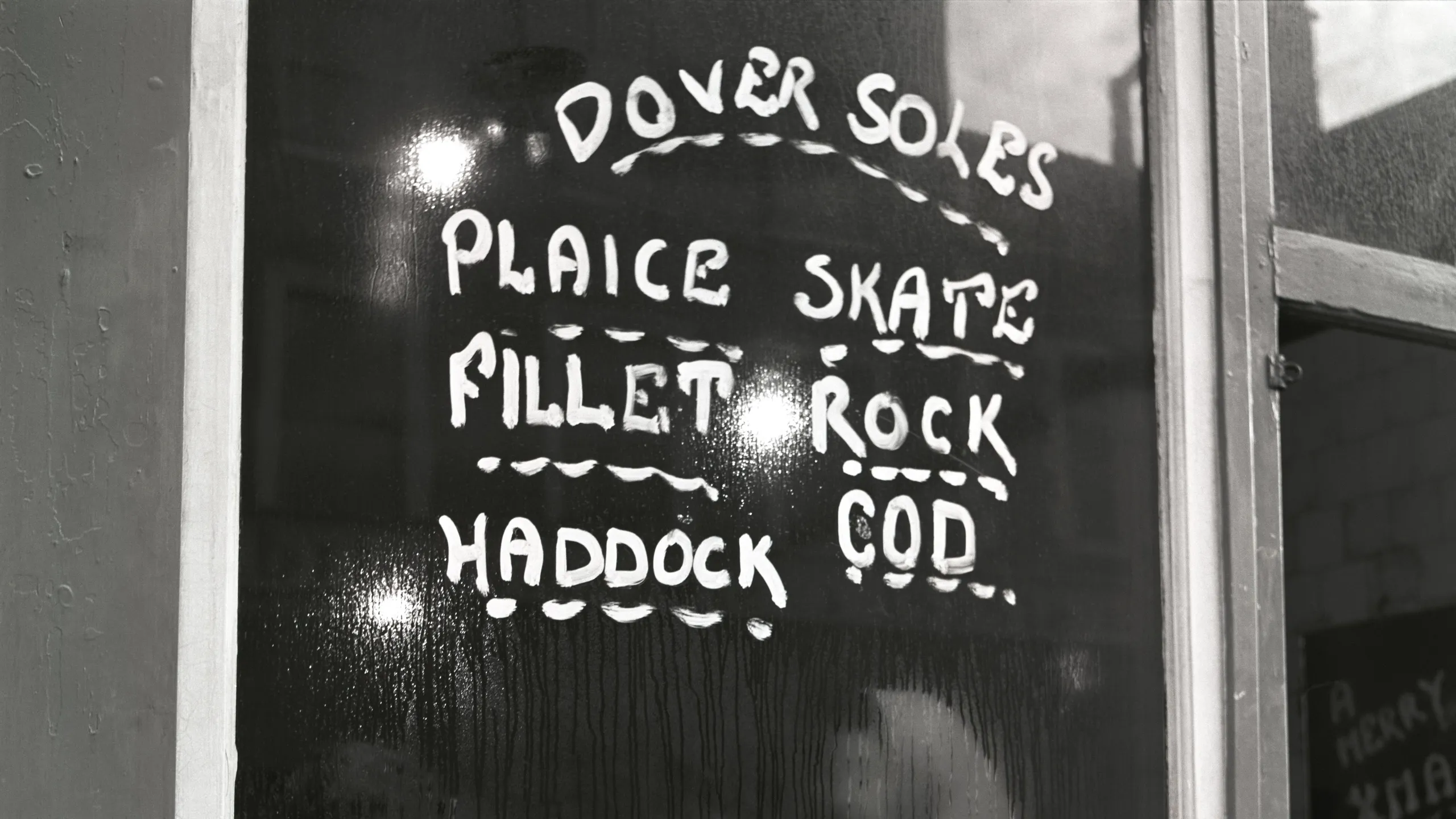 photograph, pigment print. The window of a fish and chip shop running with condensation. On it is painted the names of fish available. There is a blurred view of the interior of the shop showing jars of pickled foodstuffs, a pair of hands adding fish to the window display, and a woman in a headscarf at the counter. Through the open door to the right a stooping figure can be seen and the words 'A Merry Xmas' chalked on a board.