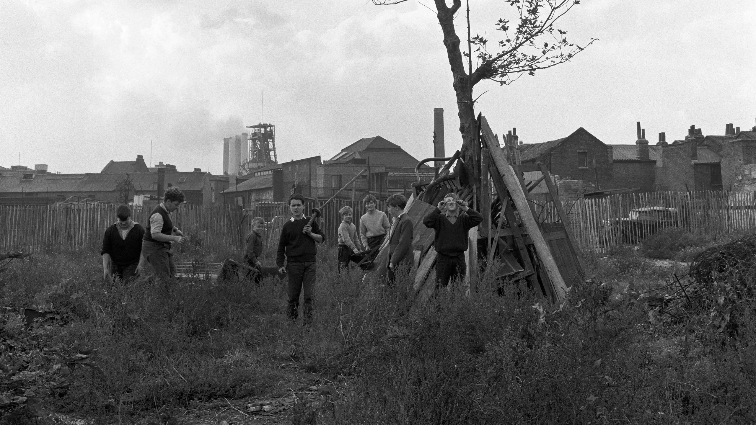 photograph, pigment print. Children playing with items that have been dumped on a patch of waste ground. Beyond the fence are houses and a power station.