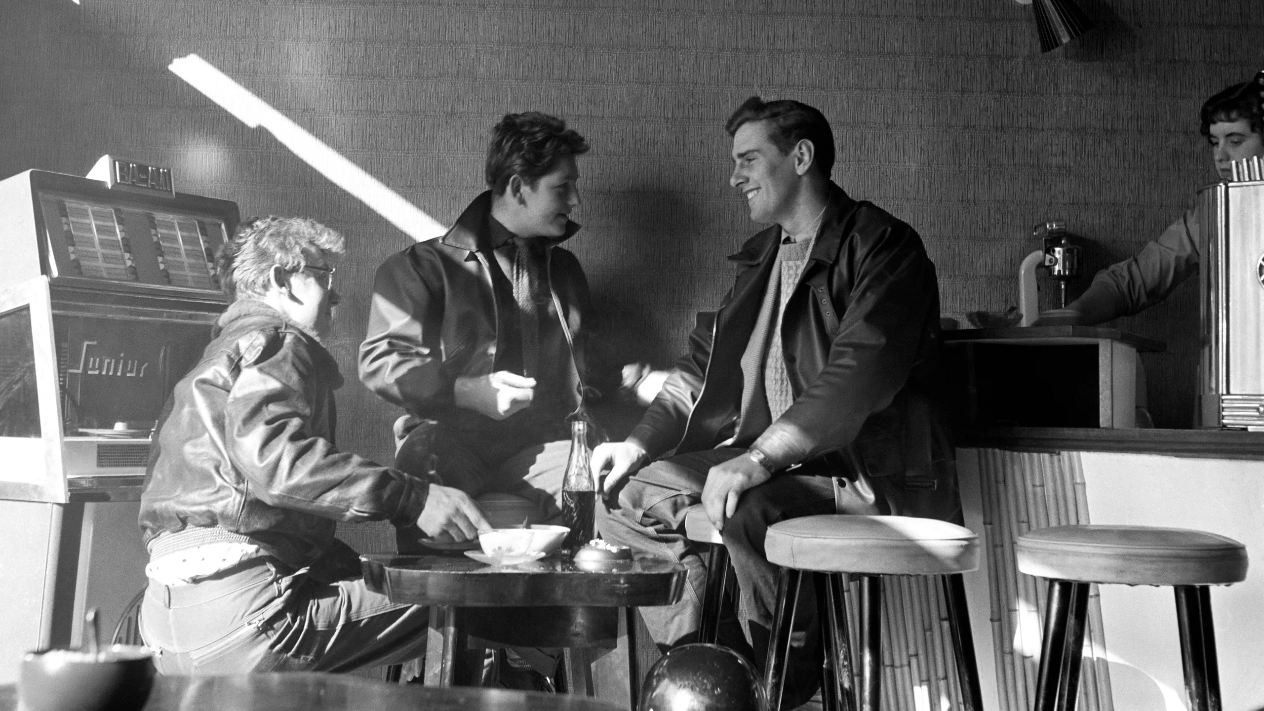 photograph, pigment print. Three young men sit around a table inside a coffee bar. The men wear jeans and zip up jackets and one has a crash helmet on the stool next to him. There is a jukebox to the left and a counter clad in bamboo to the right. A coffee machine is visible on the counter at the right edge of the image.