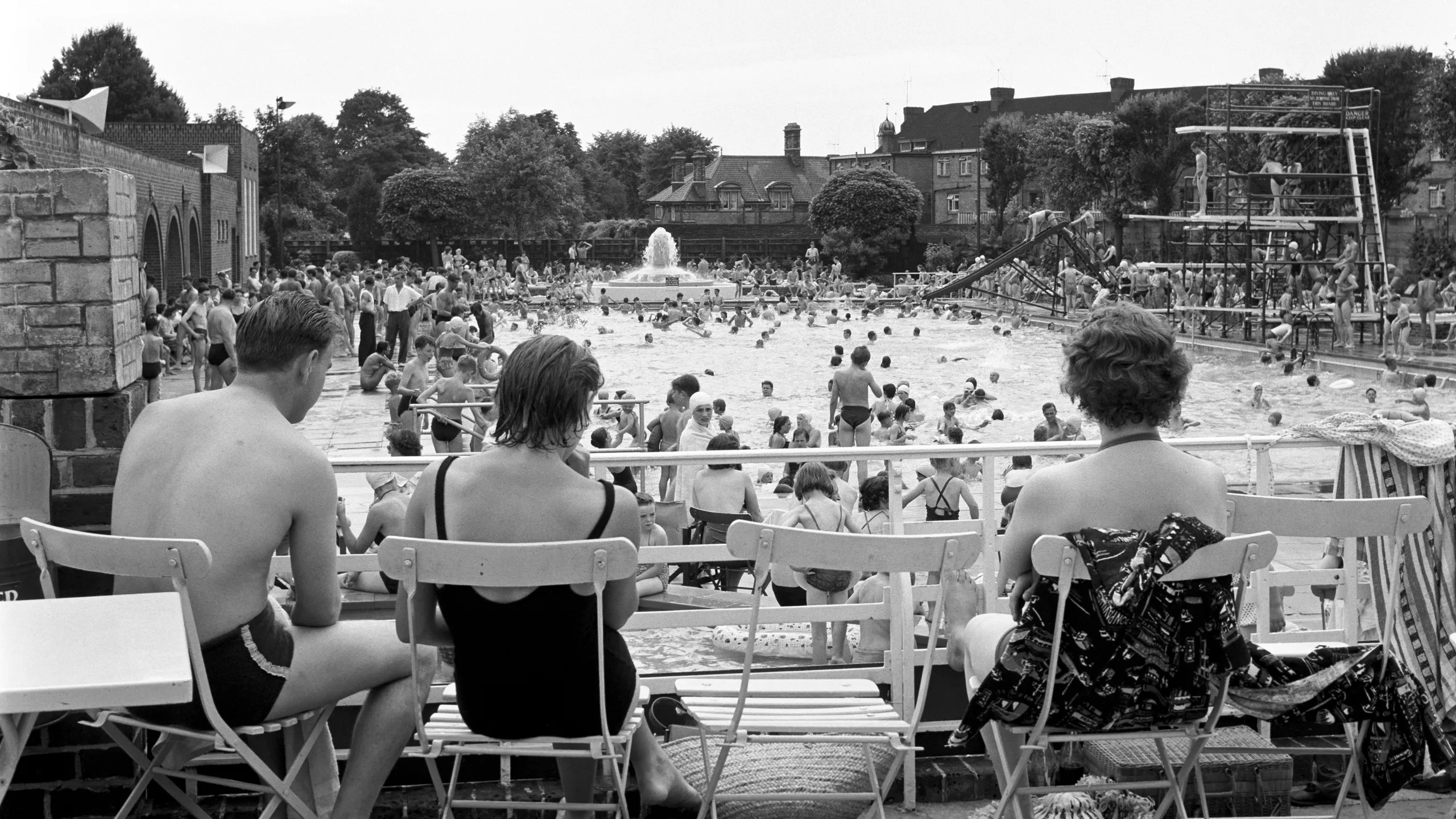 photograph, pigment print. Crowds at Twickenham Lido. People in swimming costumes surround the pool and swim in it. There is a fountain in the background and a slide and diving boards on the right of the pool. In the foreground people with their backs to the camera sit on folding chairs observing the scene.