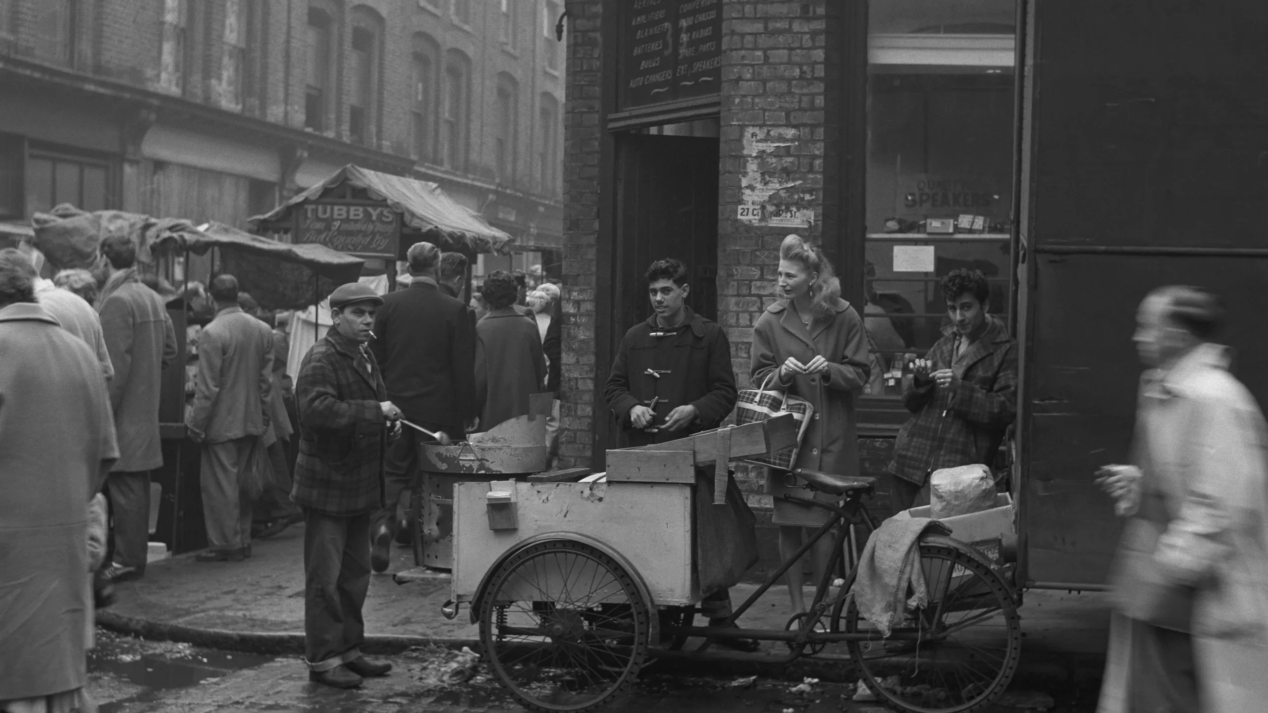 photograph, pigment print. A busy street market near Brick Lane. A man roasts chestnuts along Chilton Street in front of a shop selling electrical goods. Tubby Isaac's jellied eel stall is visible down the side street to the left.