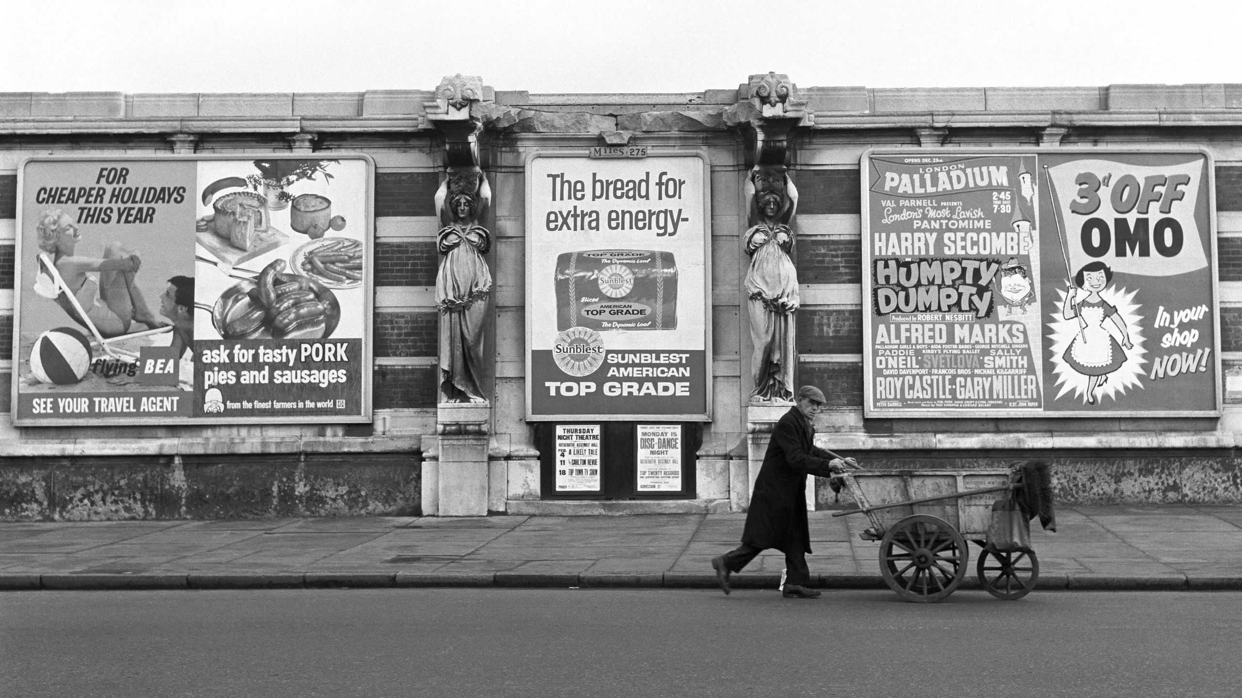 photograph, pigment print. A street sweeper wearing a coat and cap pushes his cart past a wall with two central caryatids. A row of billboards attached to the wall displays advertising posters for British European Airways, pork products, Sunblest bread, a pantomime starring Harry Secombe and Omo laundry detergent. 

Harry Secombe starred as Humpty Dumpty at the Palladium in 1959/60.