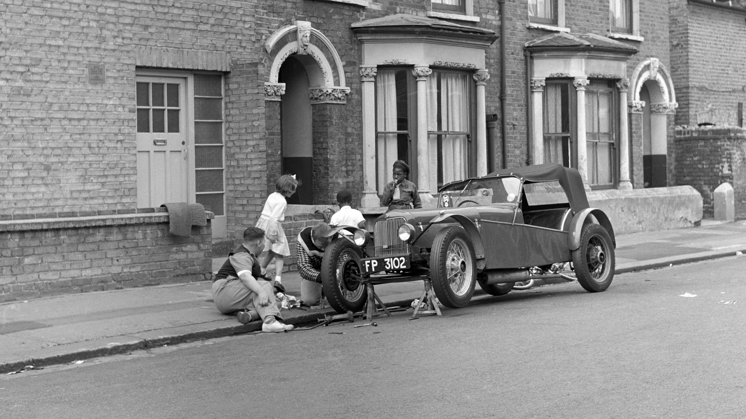 photograph, pigment print. Two men working on a car at the side of a residential street. The front of the car is supported so the tyre can be repaired. Tools lie scattered on the ground. Three children, two of them black, play on the pavement behind. The girl on the right wears a Brownie uniform.