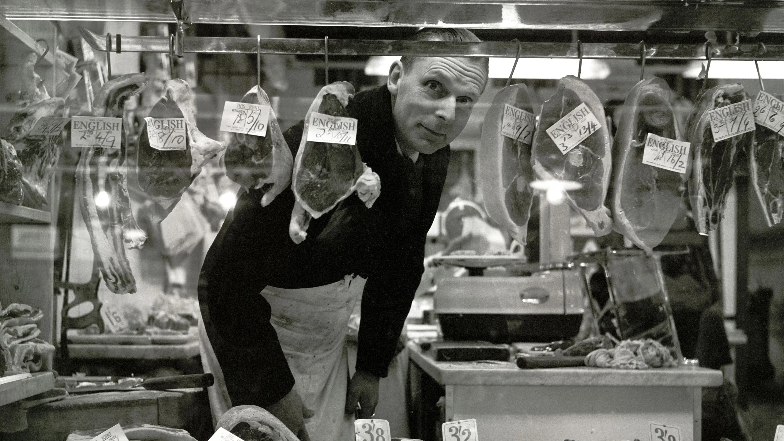 photograph, pigment print. The window display of a butcher's shop. The butcher, wearing an apron, ducks down to look at the camera through the window, so is surrounded on all sides by meat. Most of the cuts of meat are labelled 'English' together with the price per pound. Also visible through the counter is a chopping block and knife to the left and scales to the right.