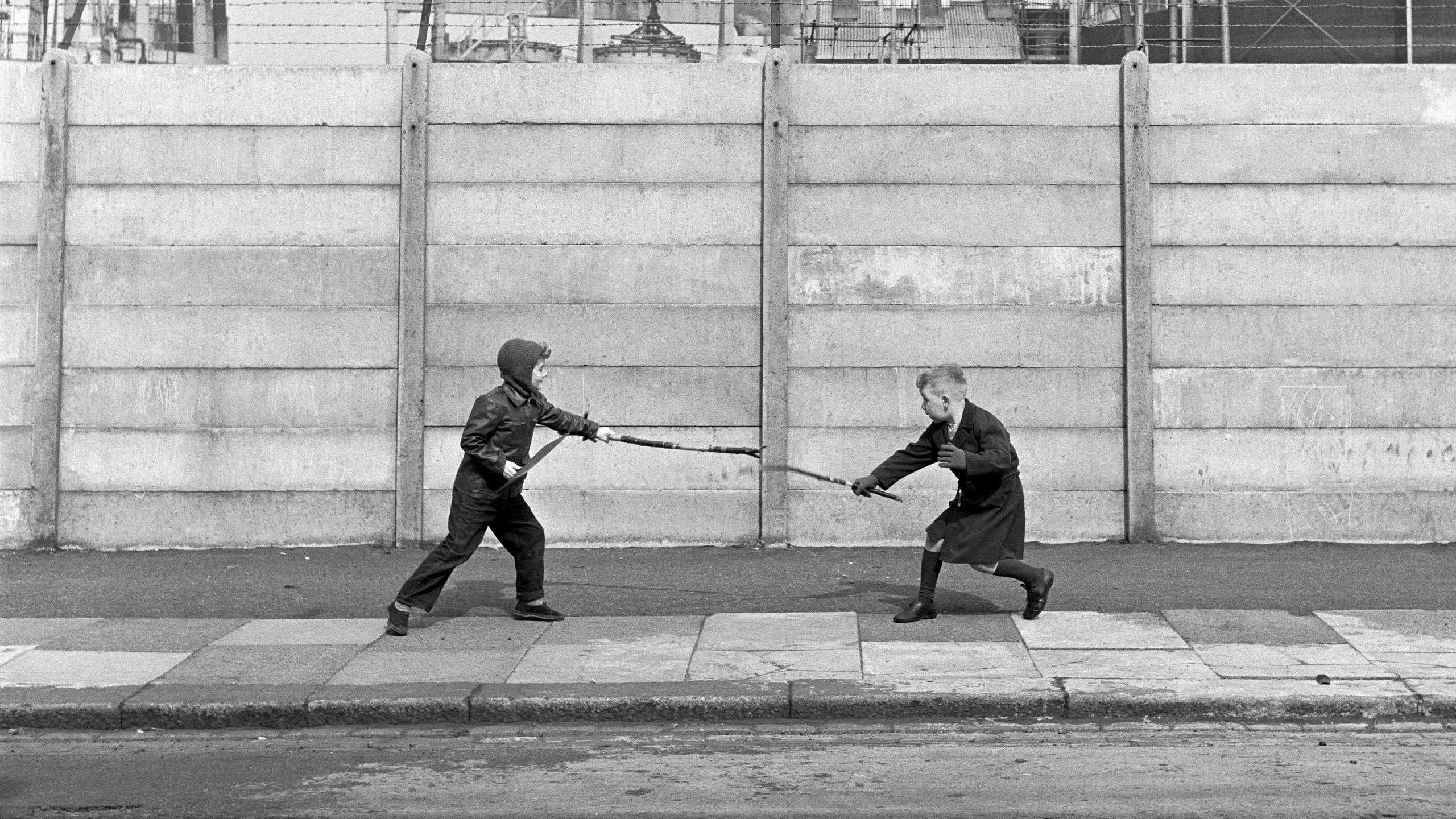 photograph, pigment print. Two boys fighting with sticks in front of a concrete wall topped with strands of barbed wire. The boy on the left holds a shield. Behind the wall is an industrial zone.