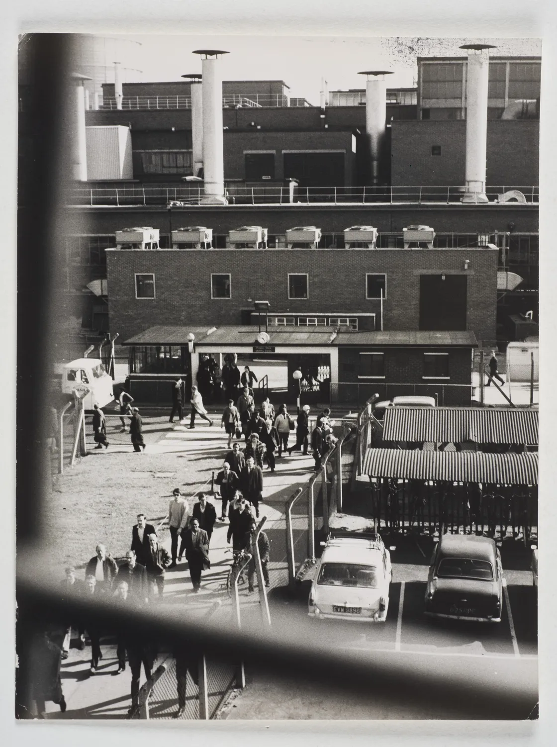 A black-and-white photo of a group of people walking through a fenced area towards a brick building with industrial machinery in the background. Several cars are parked in a nearby lot.