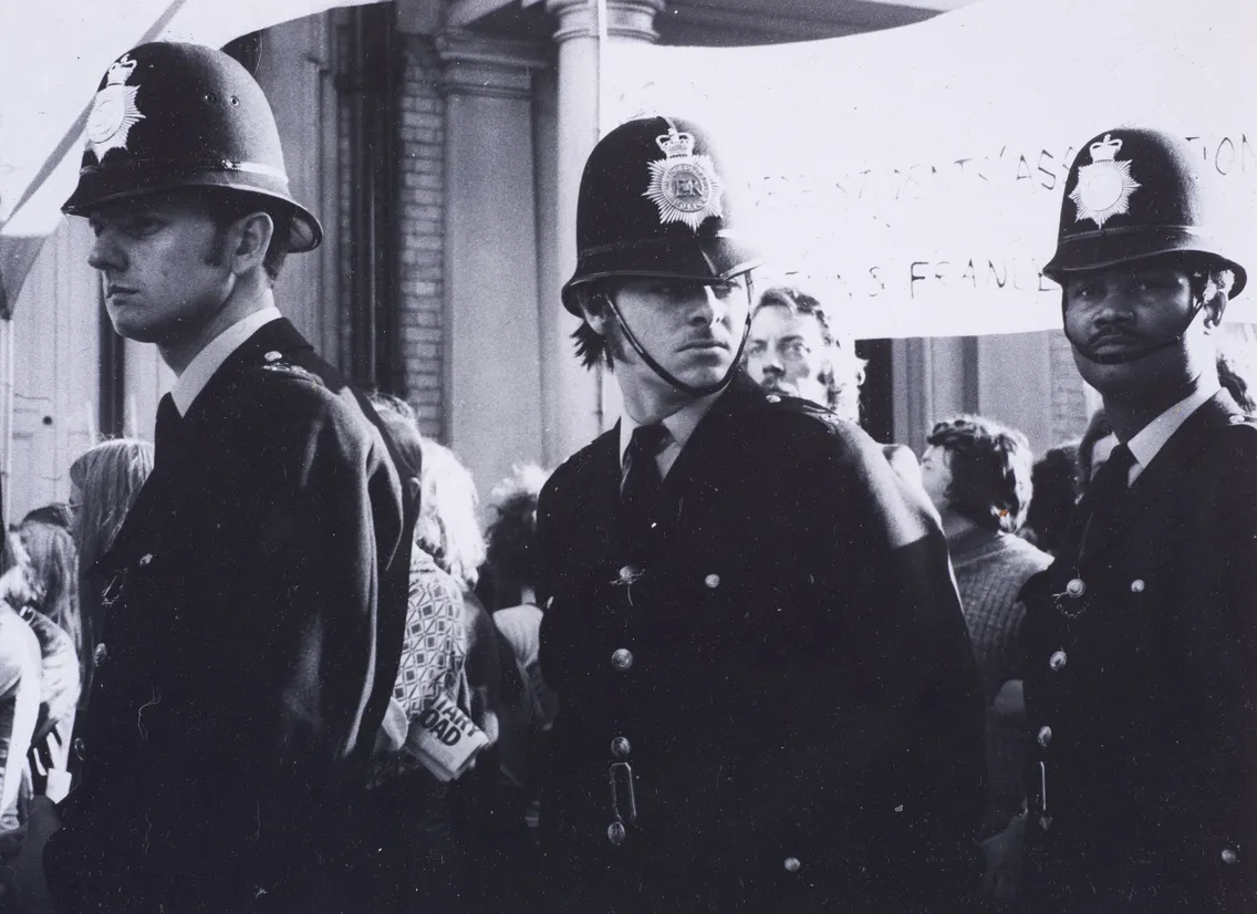 Three police officers in uniform stand in front of a crowd during a public event, with a partially visible banner in the background.