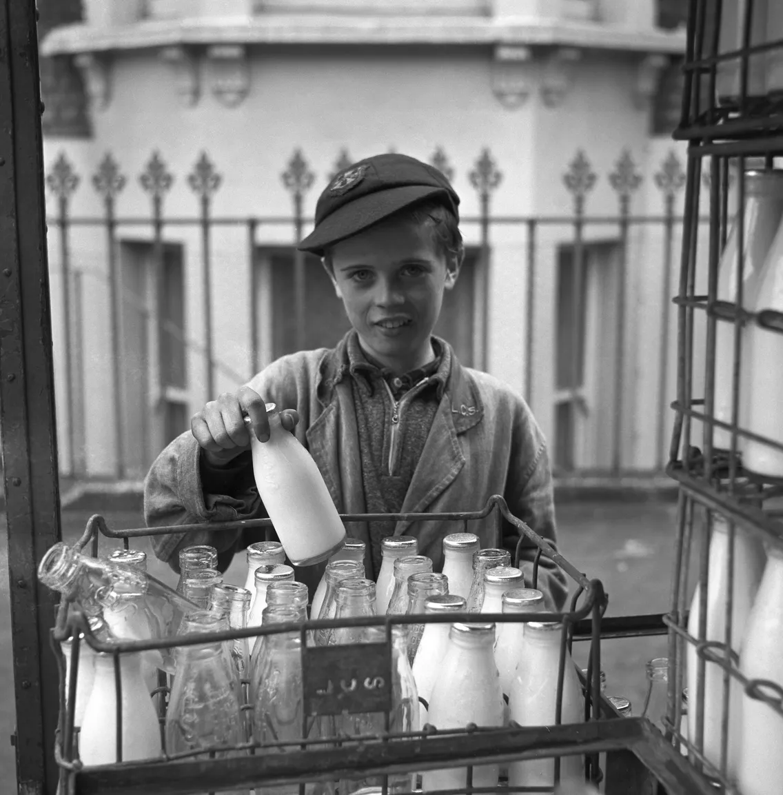 A young boy in a cap holds a milk bottle, standing behind a crate filled with empty and full milk bottles.
