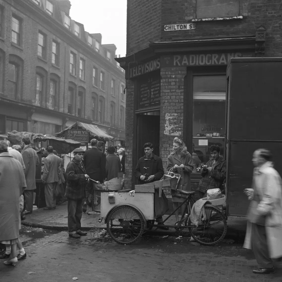 Black and white photograph of people walking and interacting around market stalls and shops in a busy urban street.
