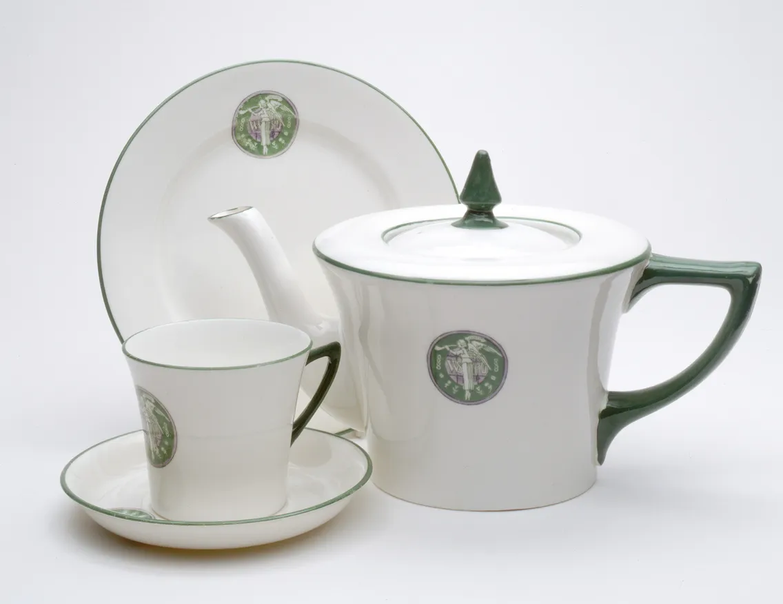 A white porcelain teapot, cup, saucer, and plate set with green accents and circular green emblems on each piece, against a plain white background.