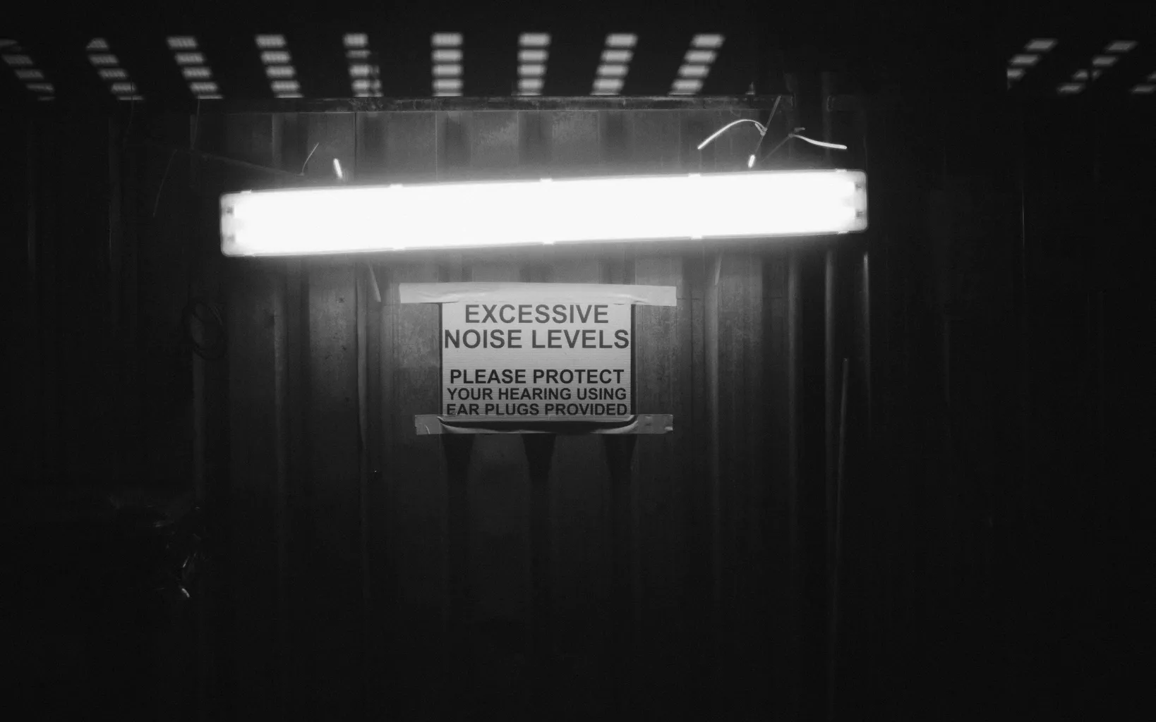 A black and white photograph of a sign reading 'Excessive noise levels. Please protect your hearing using ear plugs provided'