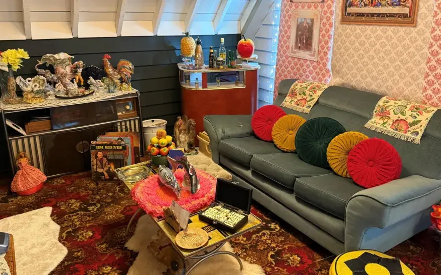 A vintage front room with a blue sofa, colorful round cushions, patterned carpet, retro decor, and various knick-knacks on shelves and tables.