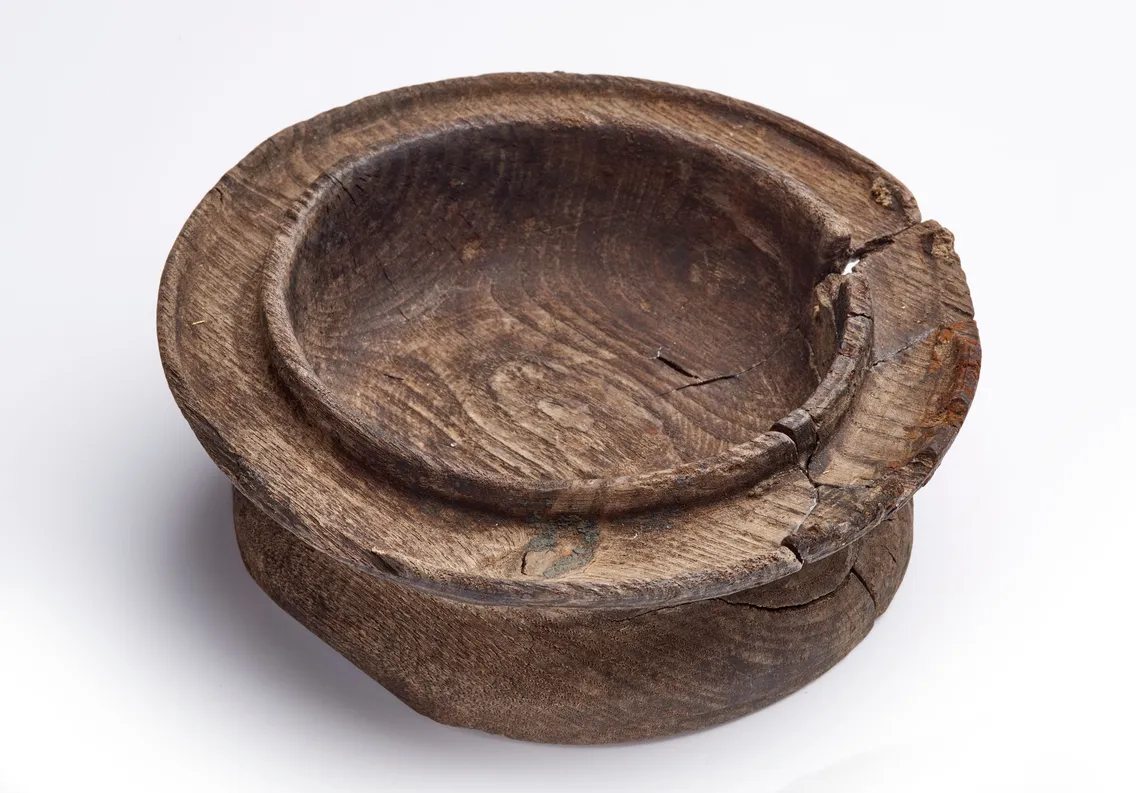 A round, shallow wooden bowl with visible cracks and a rough, weathered surface, set against a plain white background.