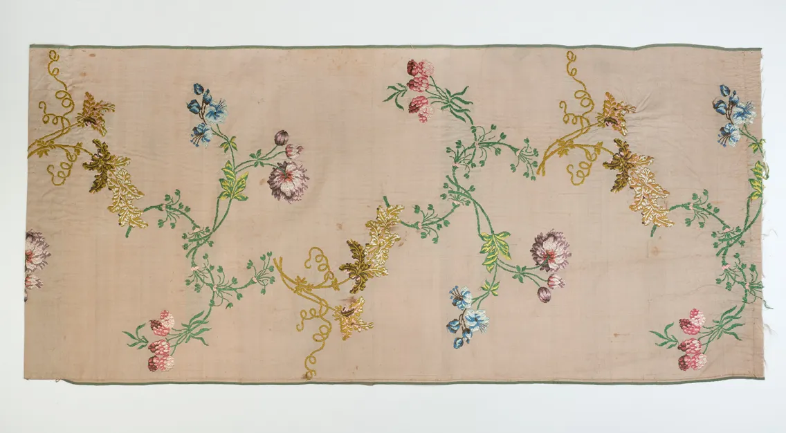 A rectangular piece of pale pink fabric featuring embroidered floral and leafy vine patterns in green, gold, red, and blue threads.