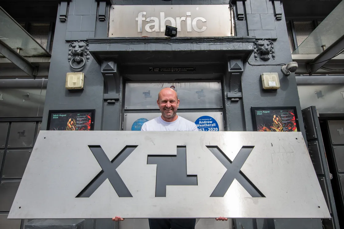 A man stands outside the entrance of a building labeled "fabric," holding a large metal sign with cut-out designs.