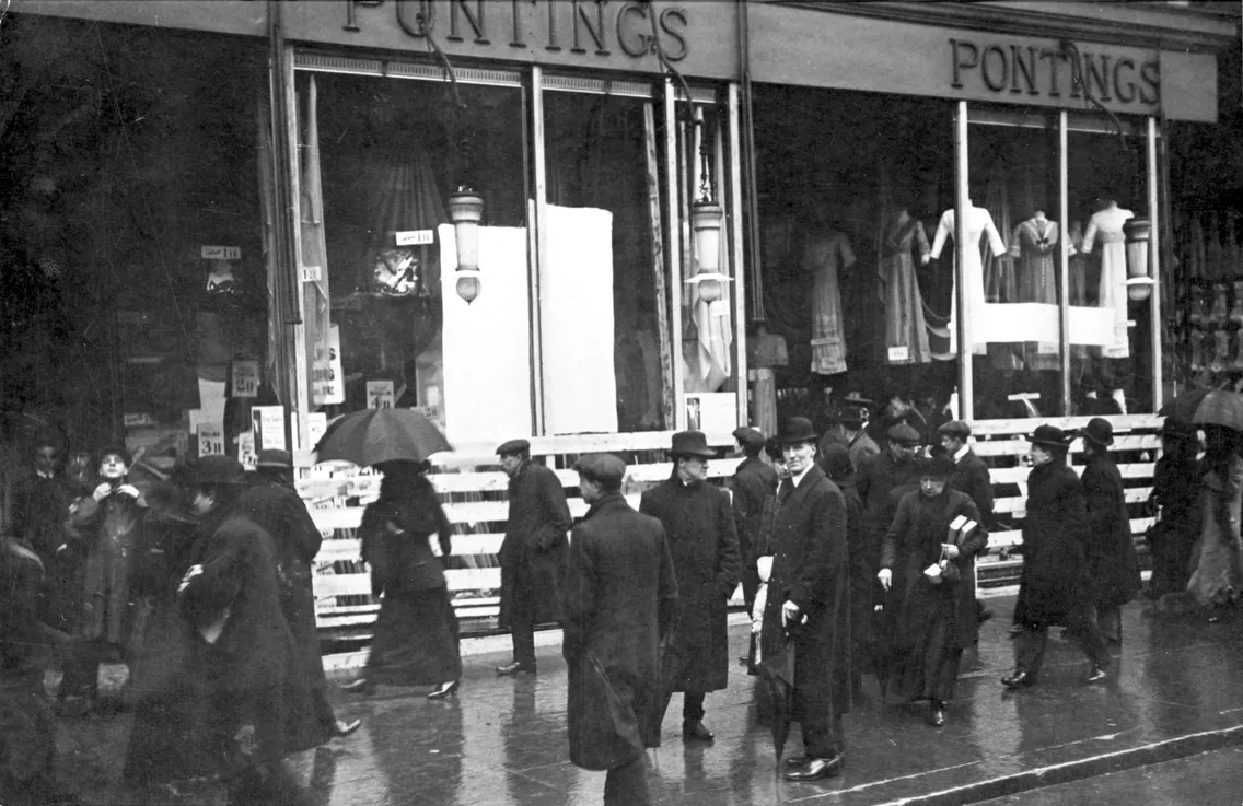 Historical black and white photo of people with umbrellas walking past pontings department store on a rainy day.