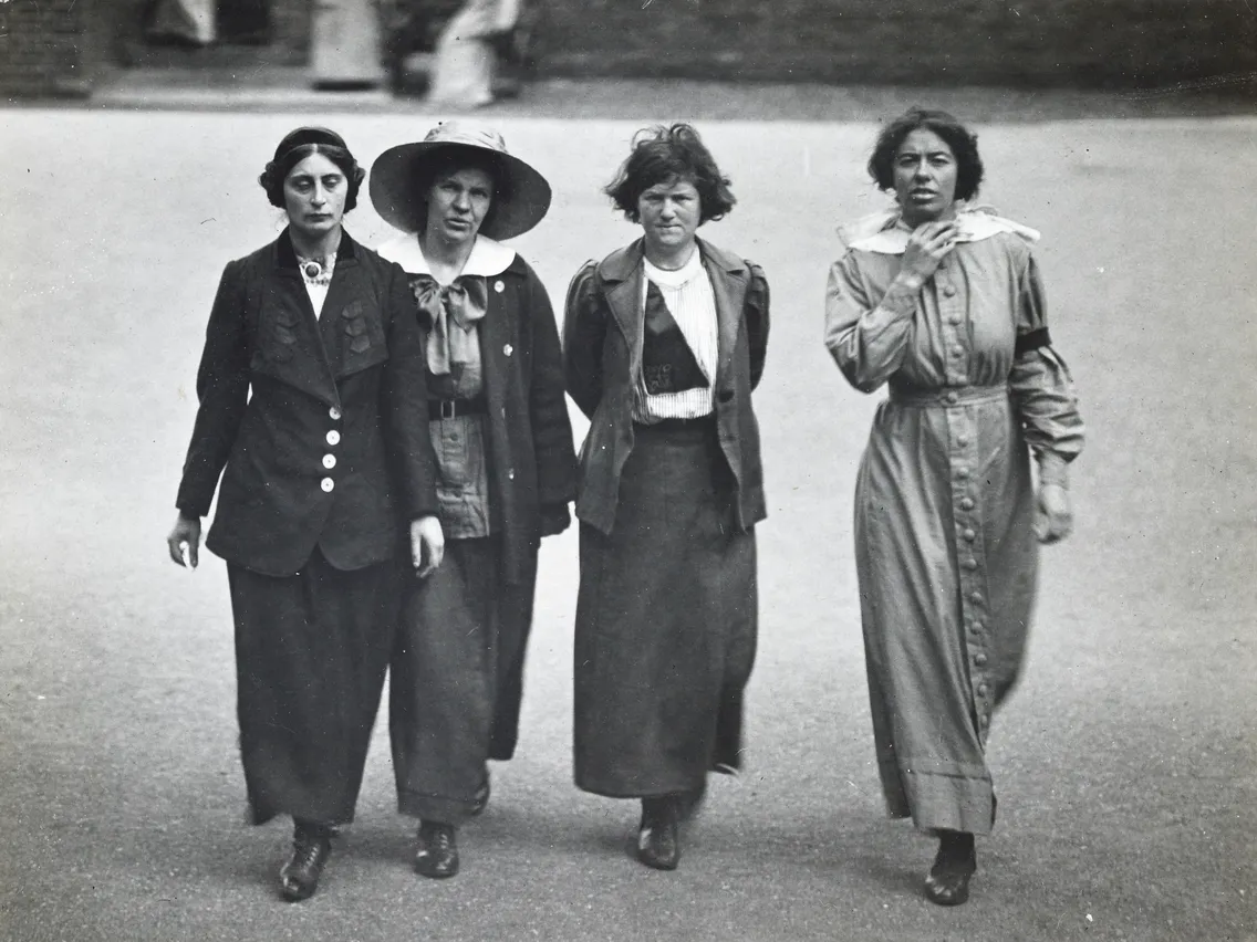 Four women from the early 20th century walking side by side on a street, dressed in period attire.