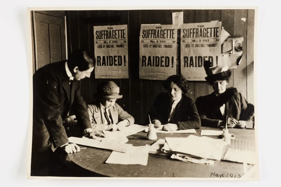 Black and white historical photo showing three women from the 20th century sitting at a table covered in papers with a figure standing beside them