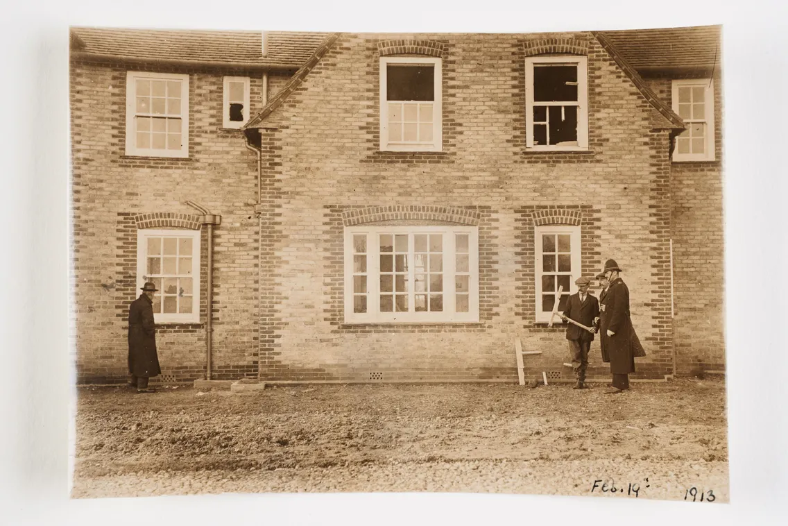 Old photograph from 1913 showing two people standing outside a brick house, one holding a framed picture.