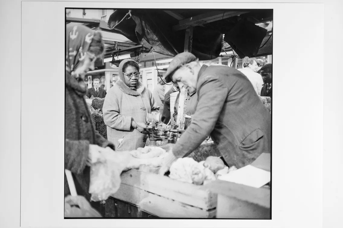A vendor weighs produce on a scale at an outdoor market stall while customers wait and observe.