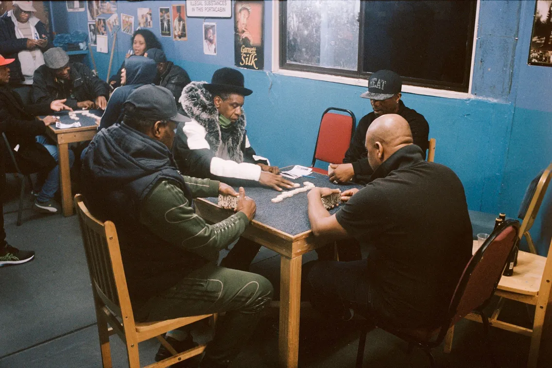 Four people are seated around a table inside a room, focused on playing a game of dominos. Other people are seen in the background engaged in a similar activity.