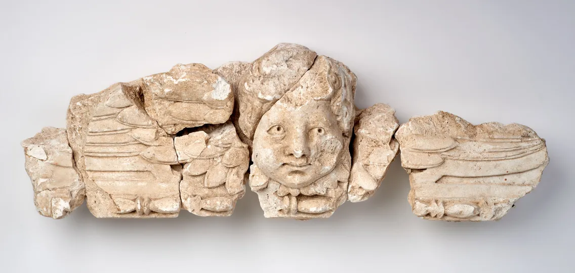 Fragmented stone relief depicting a central human face with stylized wings or drapery extending on both sides.