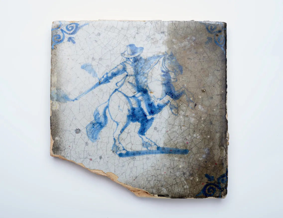 A ceramic tile with a blue illustration of a person on horseback, wearing a hat and holding a long object, with decorative blue corners and a chipped edge.